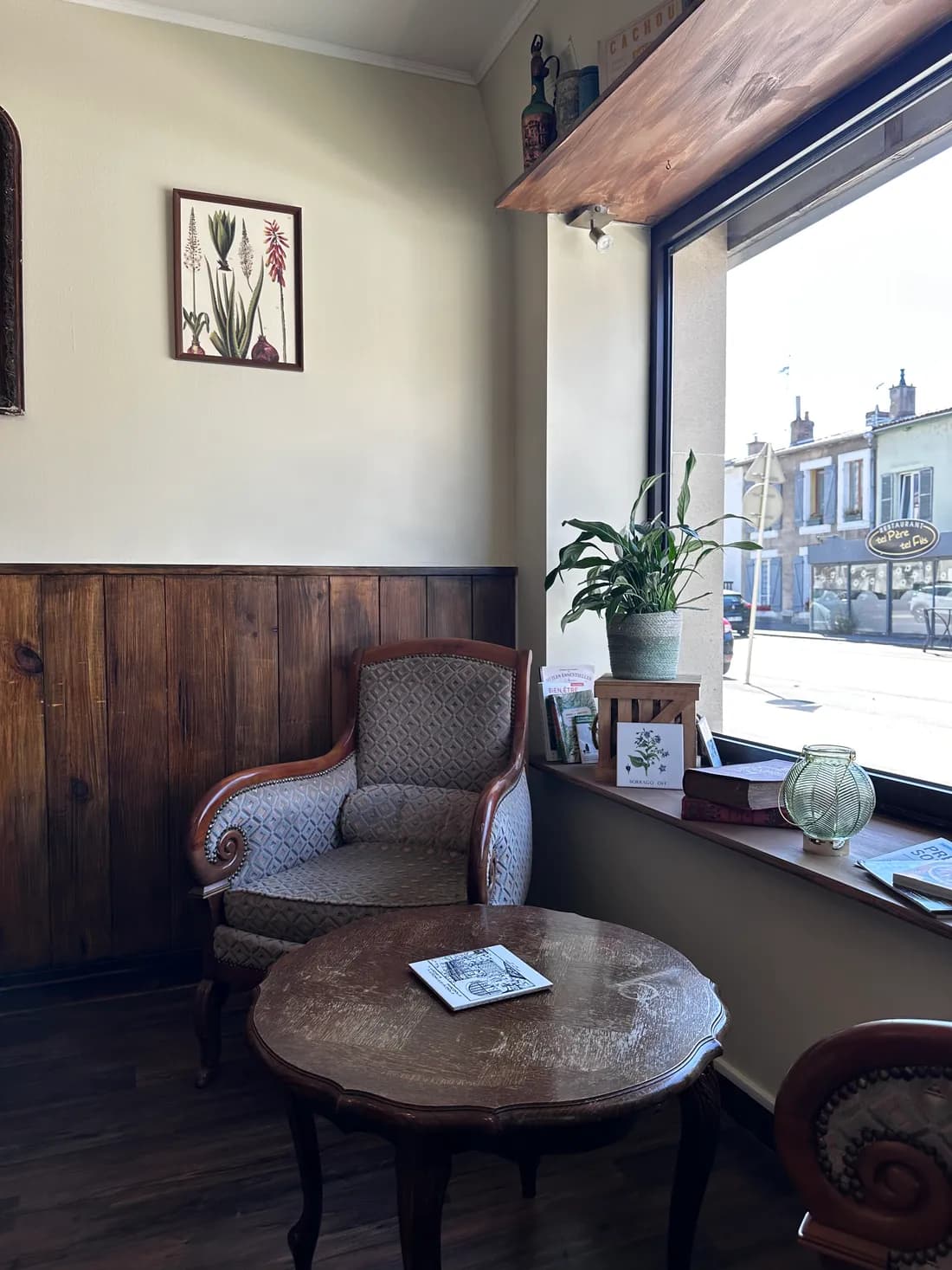 Le coin lecture du Bistrot : fauteuil ancien, table basse et planche botanique, devant la fenêtre sur la rue